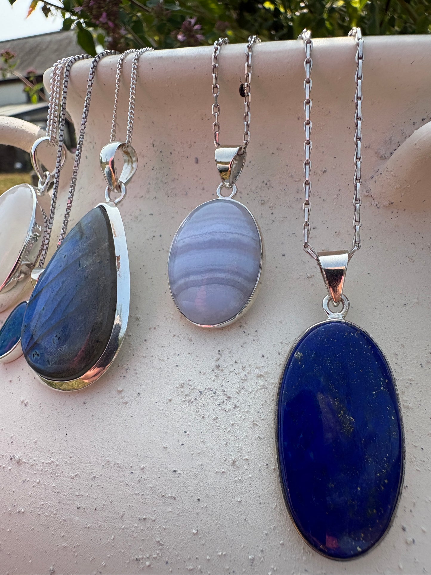 Three silver chain necklaces with oval and teardrop-shaped gemstone pendants displayed on a light beige textured surface. The pendants include a deep blue stone, a lavender and white banded stone, and a dark blue-gray stone.