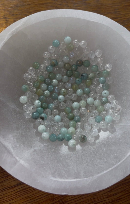 Amazonite and Rainbow Moonstone beads resting in a carved selenite bowl before being handcrafted into a sacred geometric tantric necklace, photographed in natural light.