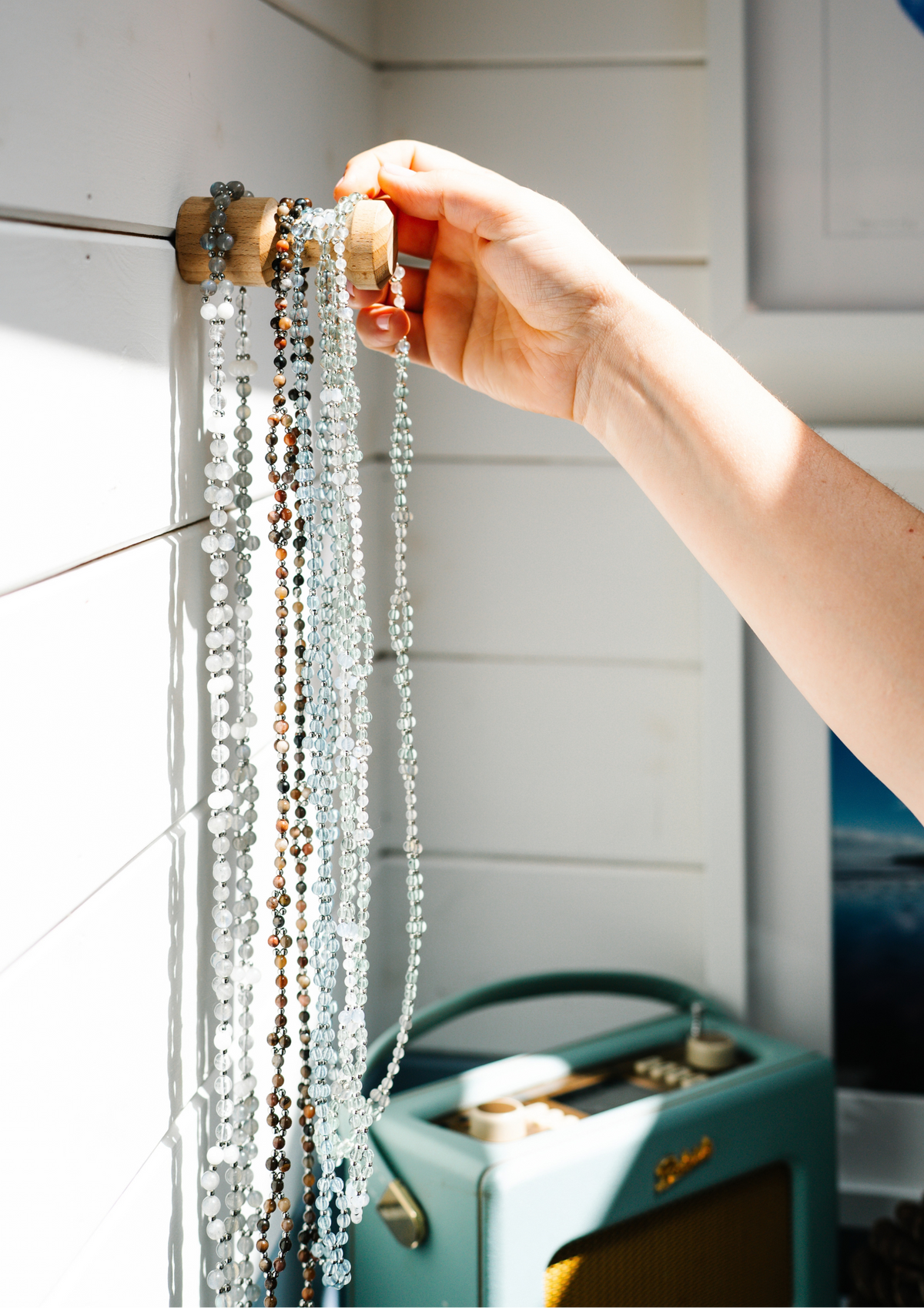 A hand holding a clear beaded necklace against a white shiplap wall, with a turquoise vintage radio and framed art in the background.