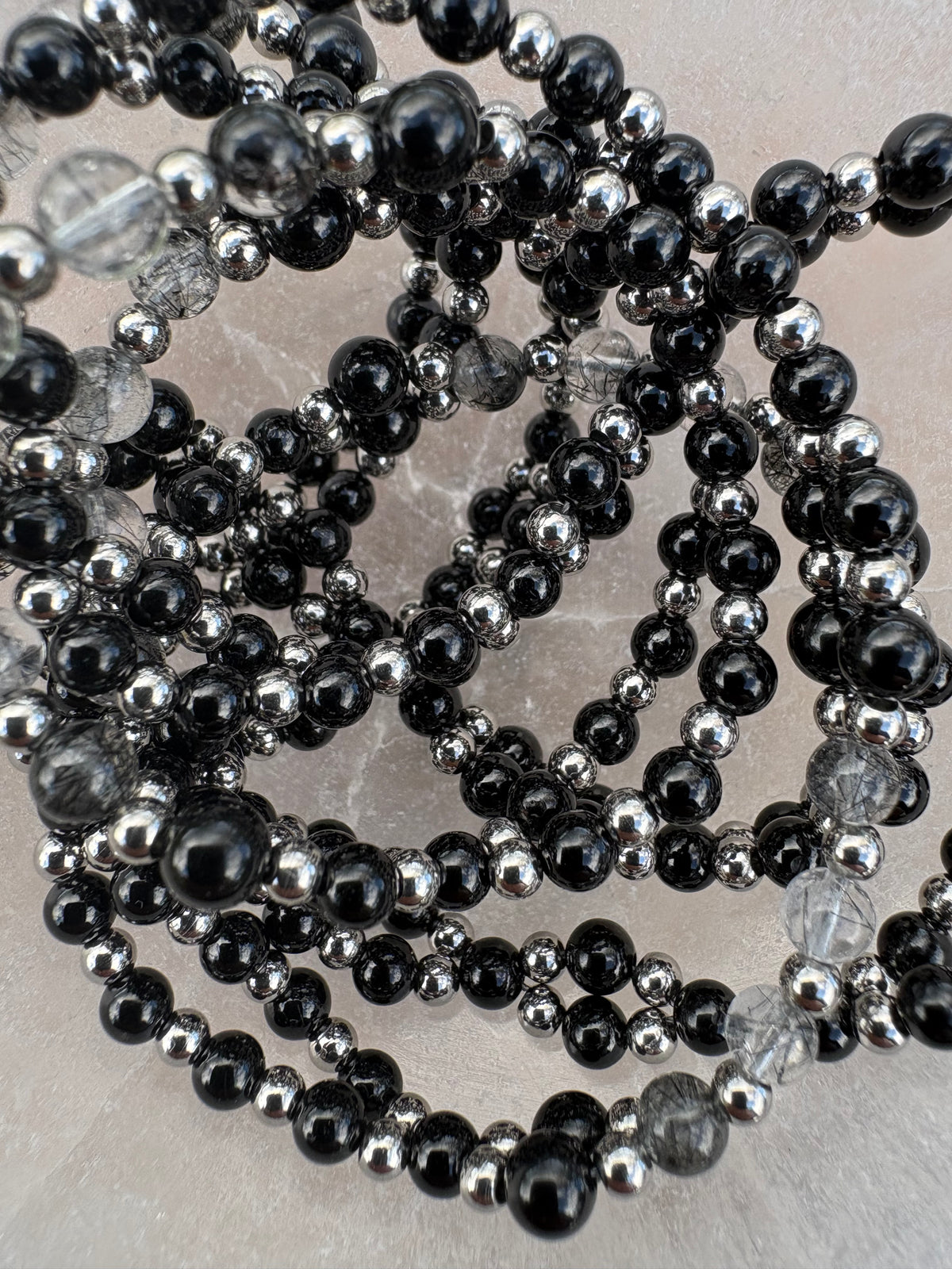 Close-up of a beaded strand featuring alternating glossy black and polished silver round beads, with a few clear faceted beads visible, arranged on a light neutral surface.