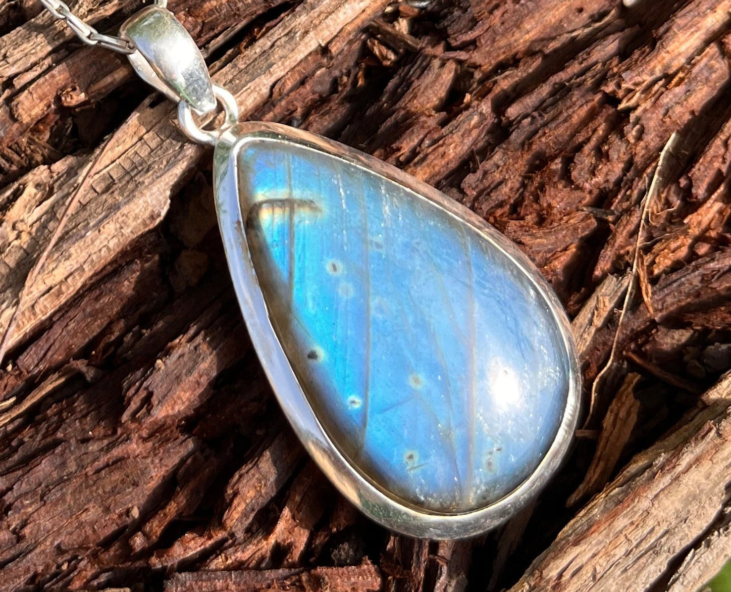 A teardrop-shaped pendant necklace with a shimmering blue iridescent stone in a silver bezel setting, resting on a textured piece of weathered wood.