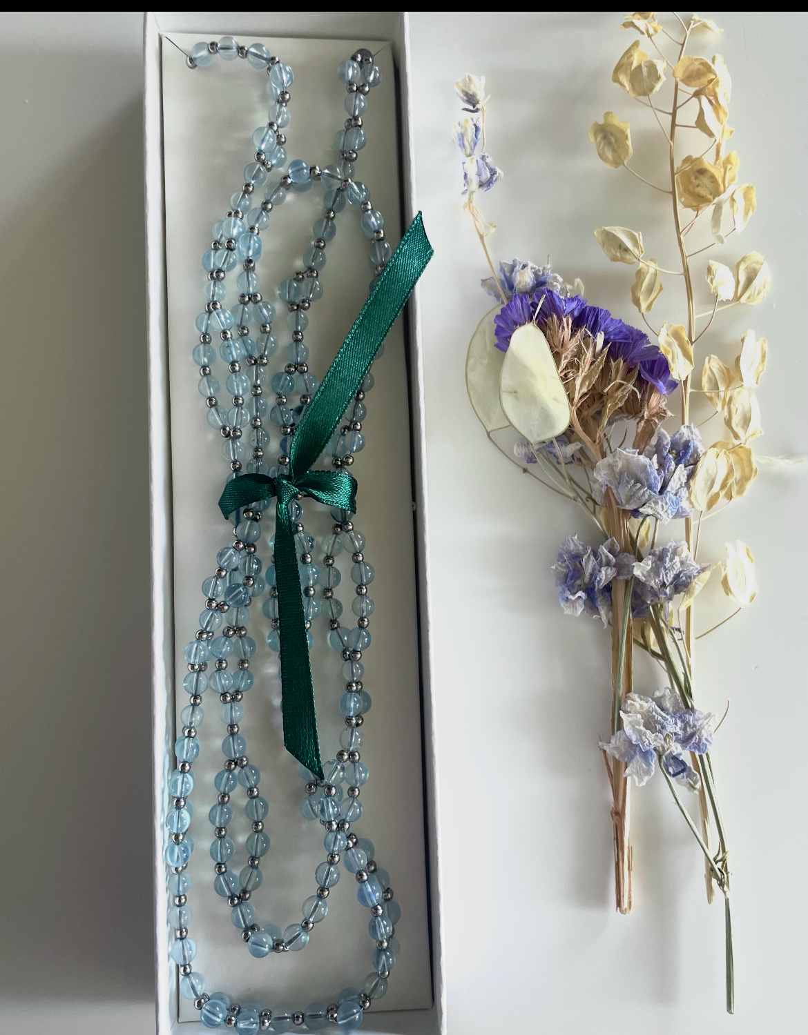 A long beaded necklace with light blue beads and a dark green ribbon bow, displayed next to a small dried flower bouquet on a white surface.