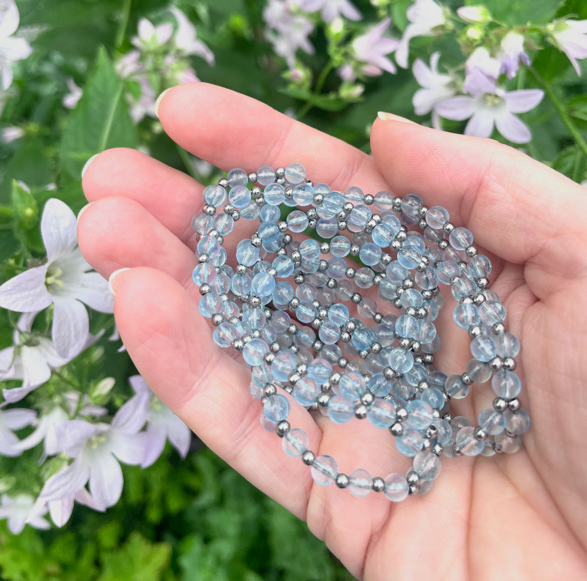 A hand holding a coiled bundle of light blue-gray rhinestone beads or chains, photographed outdoors against a blurred background of green leaves and small white flowers.