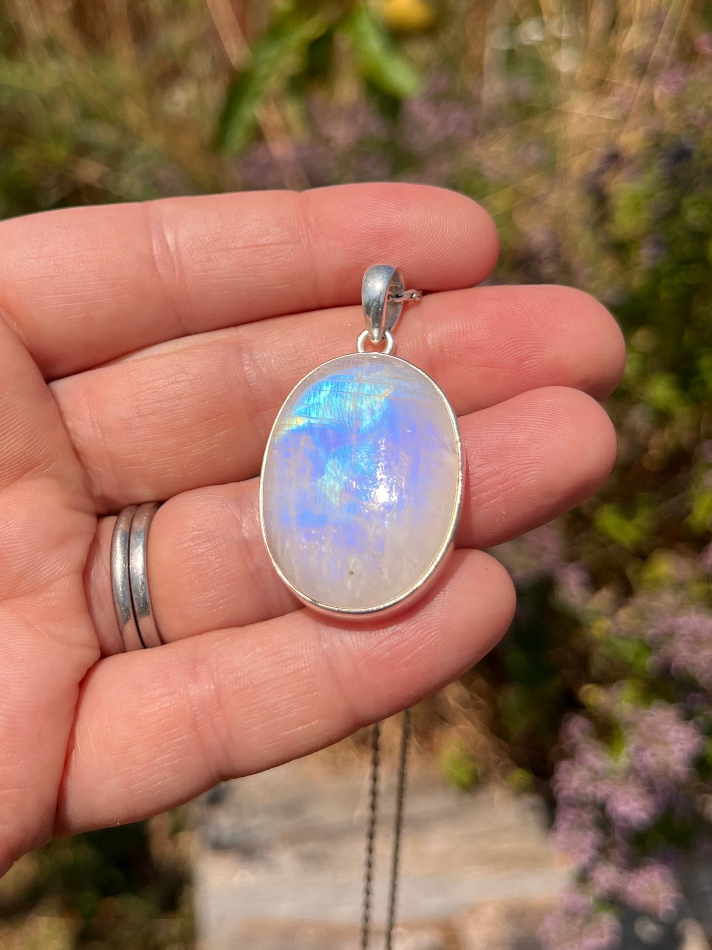 A silver pendant necklace featuring an oval cabochon moonstone with iridescent blue, white, and purple flashes, held in a person's hand against a blurred outdoor background of greenery and flowers.