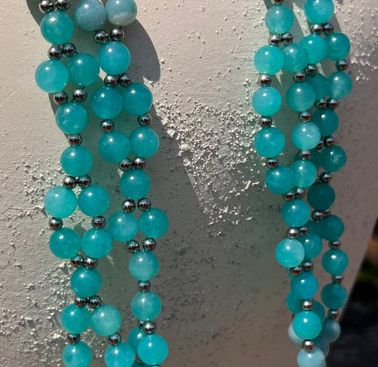 A beaded strand necklace with turquoise-blue and white beads draped over a white ceramic pitcher outdoors on a wooden surface.