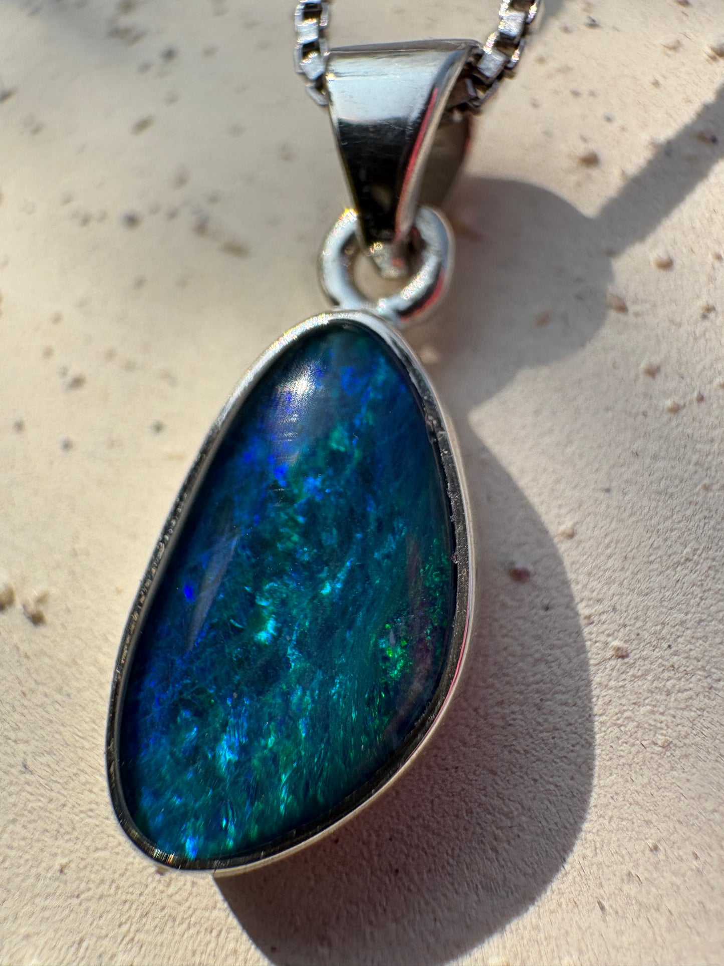 A close-up of a teardrop-shaped pendant with a silver bezel setting and a highly iridescent blue opal inlay, resting on a textured beige surface.