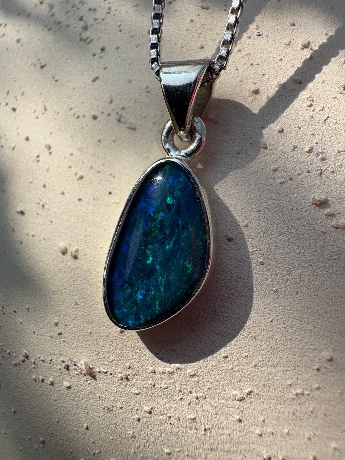A teardrop-shaped pendant necklace featuring a vibrant blue and green iridescent opal stone in a silver bezel setting, resting on a textured beige surface.