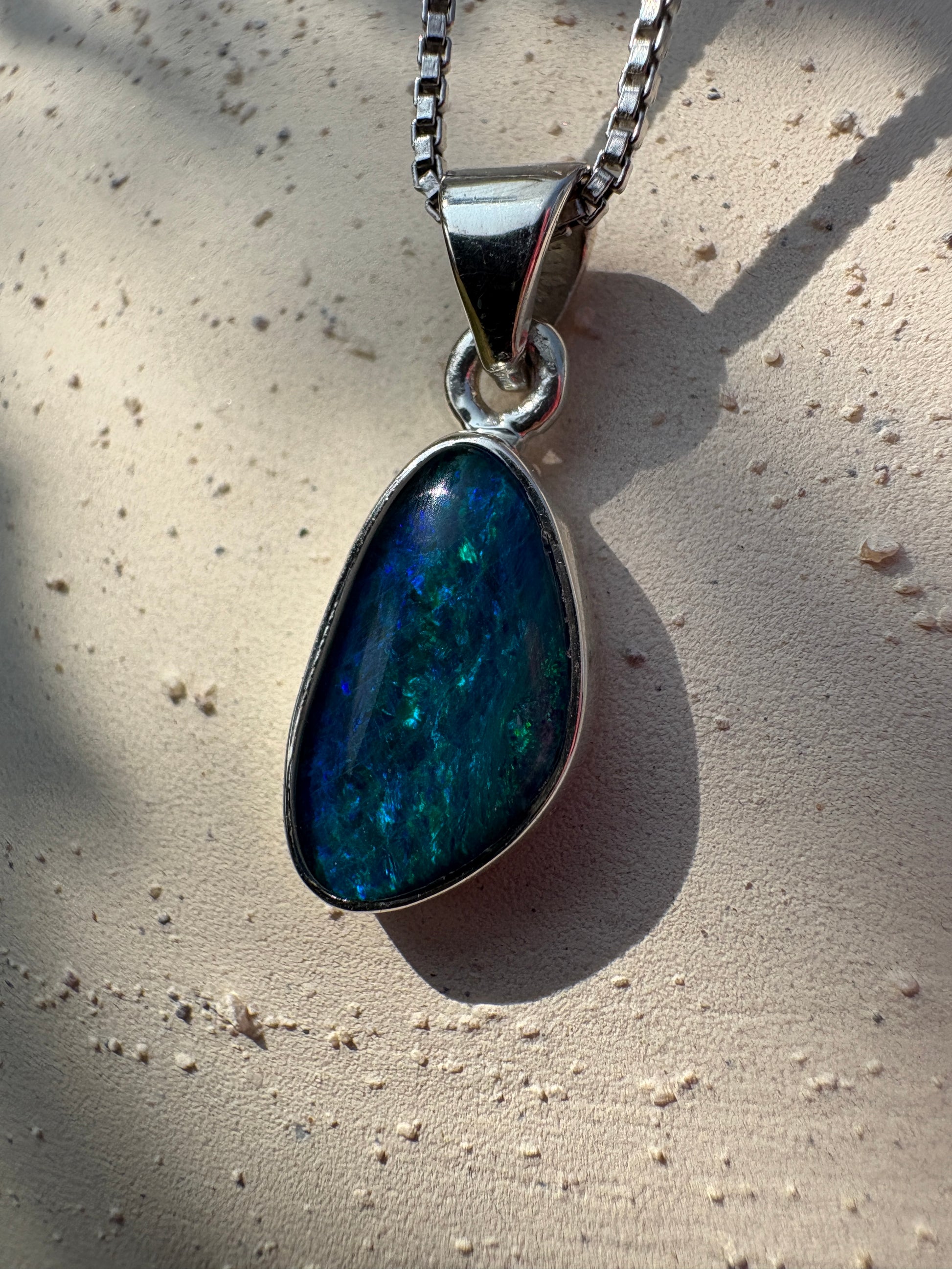 A teardrop-shaped pendant necklace featuring a vibrant blue and green iridescent opal stone in a silver bezel setting, resting on a textured beige surface.