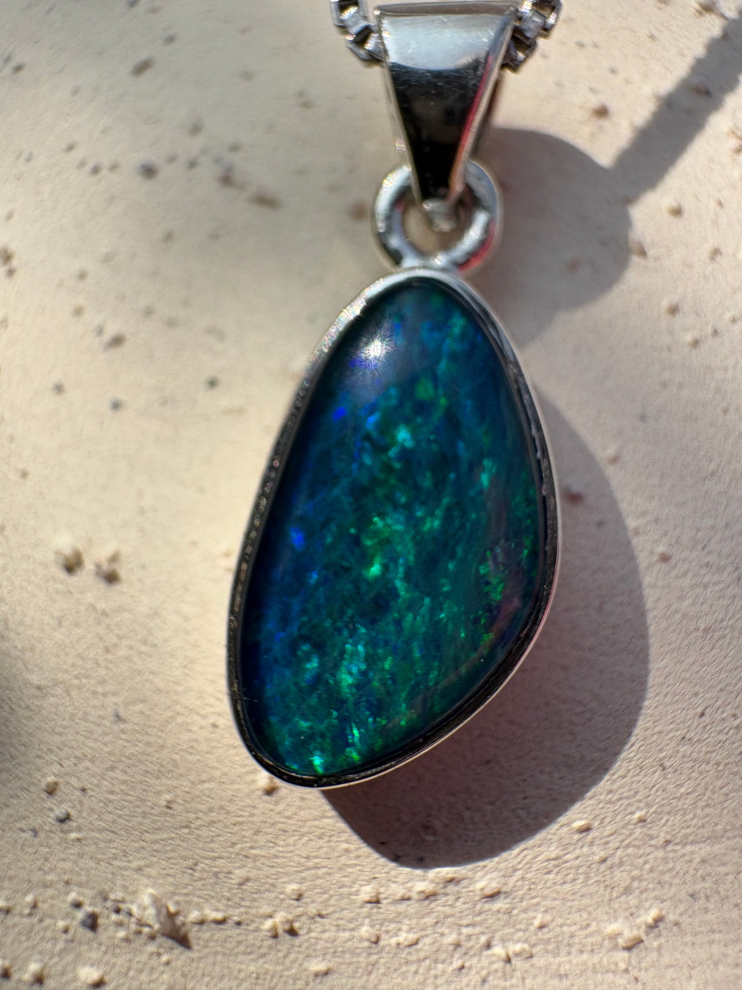 A close-up of a teardrop-shaped pendant with a silver bezel holding a vibrant blue and green opal, shown on a textured beige surface with a hard shadow.