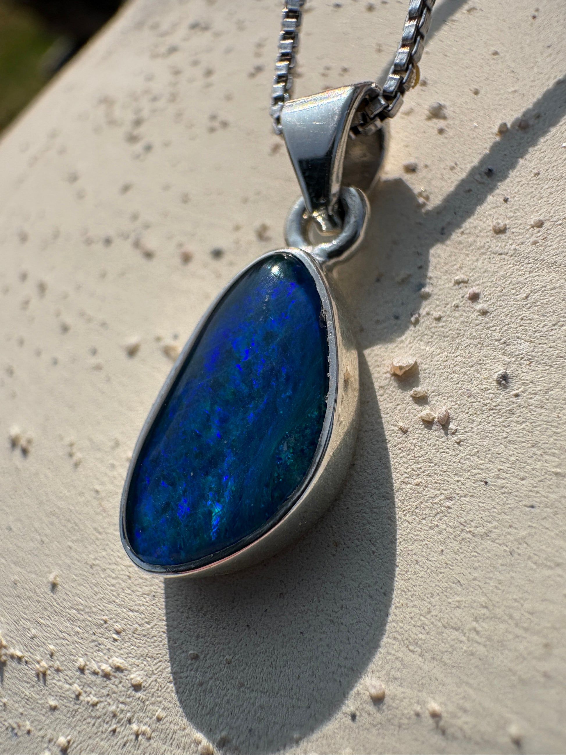 A silver pendant necklace with a teardrop-shaped blue opal stone, resting on a textured beige stone surface.