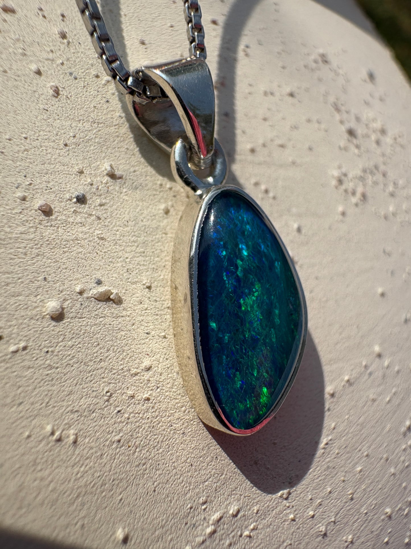 A silver teardrop pendant necklace featuring a vibrant blue and green iridescent opal stone, resting on a textured light beige surface.