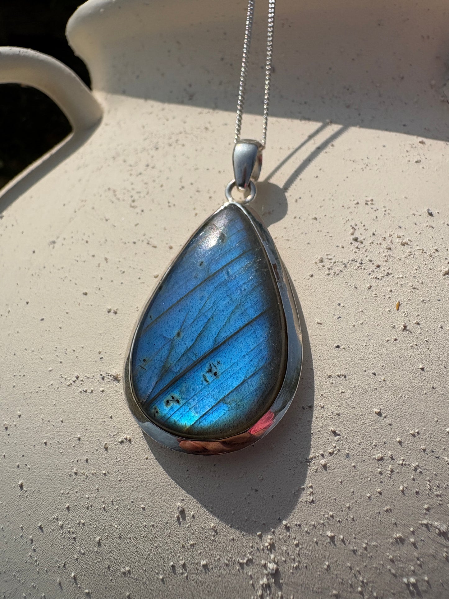 A teardrop-shaped pendant necklace featuring a blue iridescent labradorite stone in a silver bezel setting, hanging from a silver chain against a textured off-white background.
