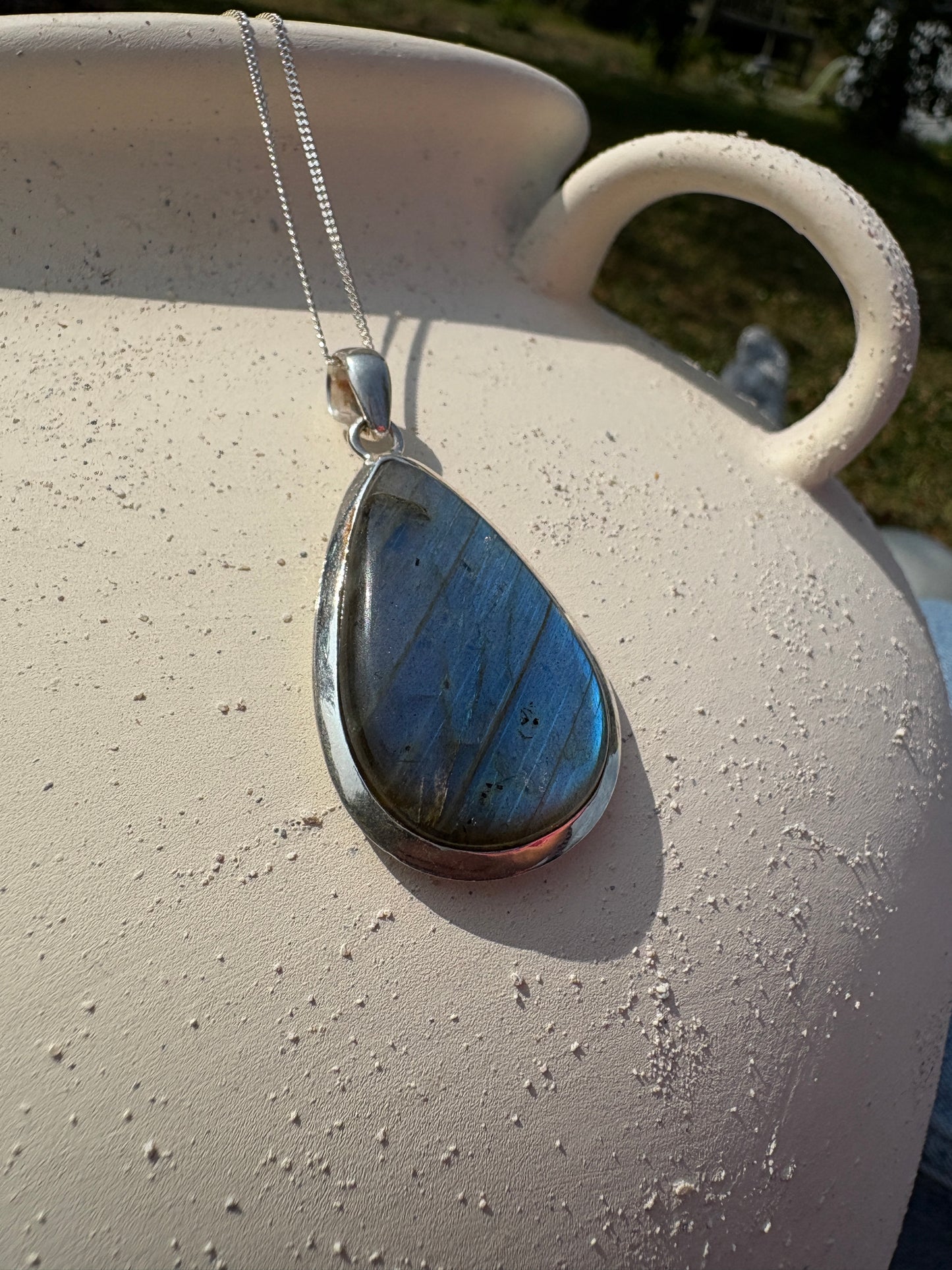 A teardrop-shaped labradorite pendant necklace with a silver bezel and chain, resting on a textured beige ceramic pot outdoors.