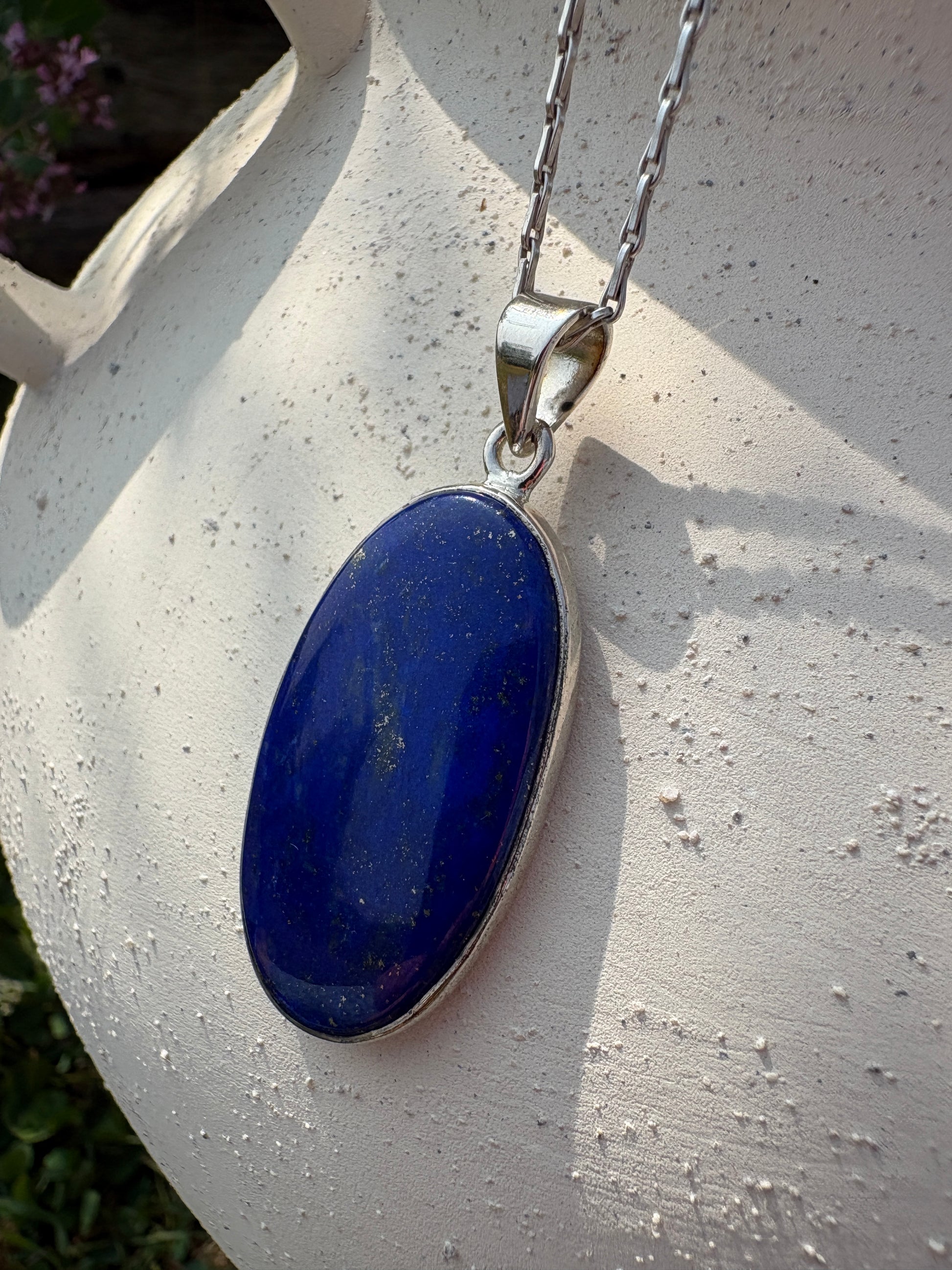 A silver oval pendant necklace with a deep blue lapis lazuli stone, resting on a textured white surface.