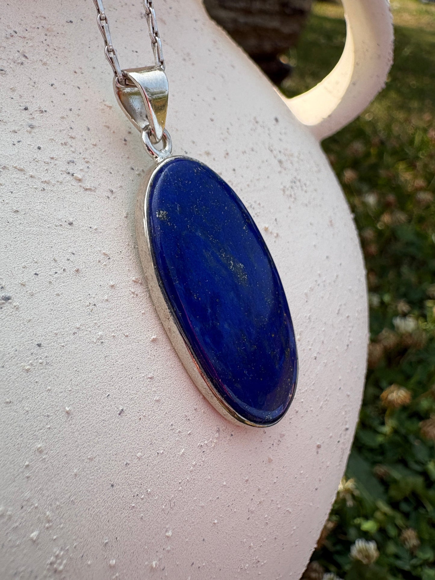 A silver necklace with an oval dark blue lapis lazuli pendant, resting on a textured white ceramic surface with a blurred garden background.