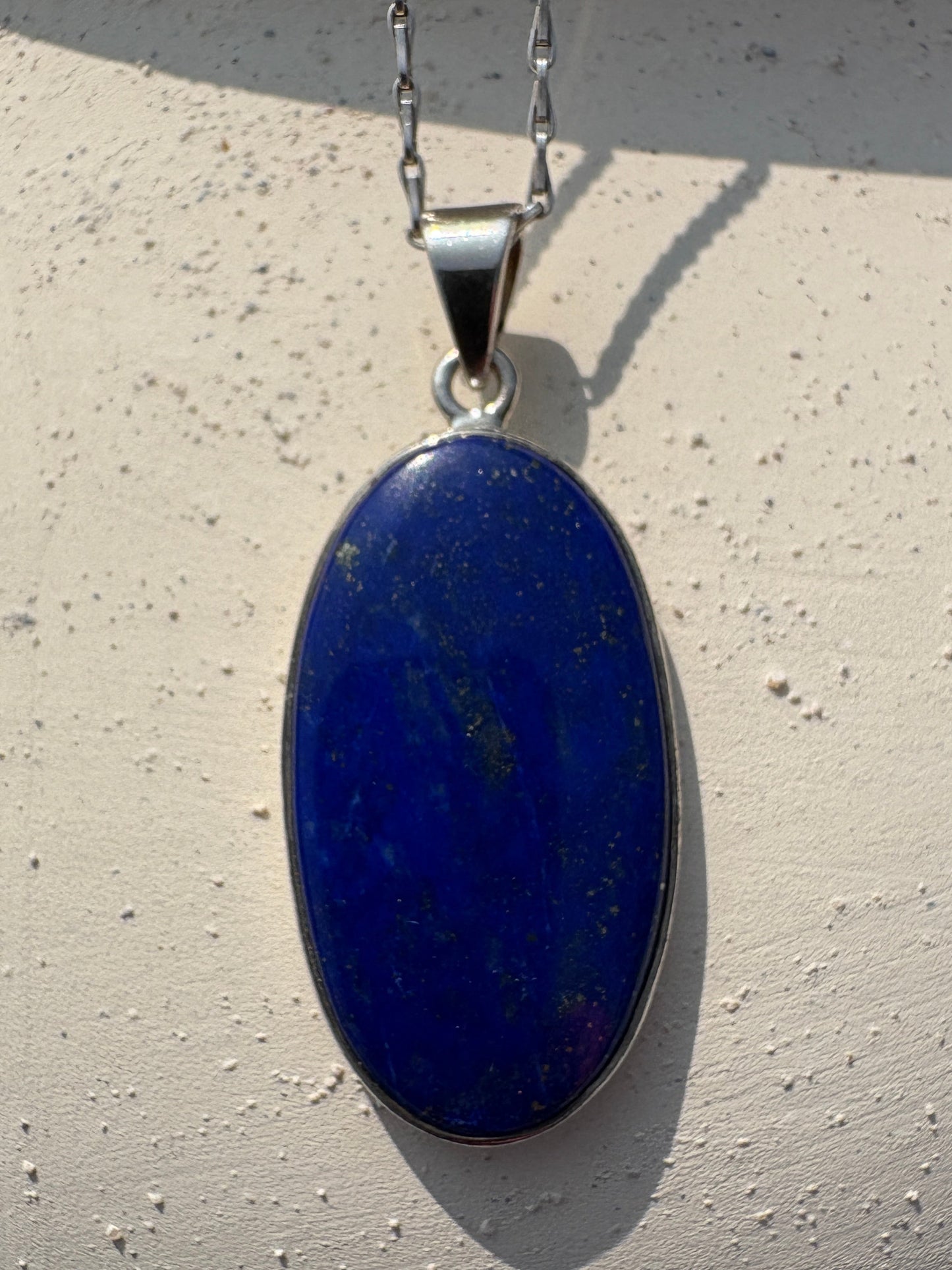 An oval-shaped dark blue lapis lazuli pendant necklace with a silver bezel and chain, resting on a textured off-white surface.