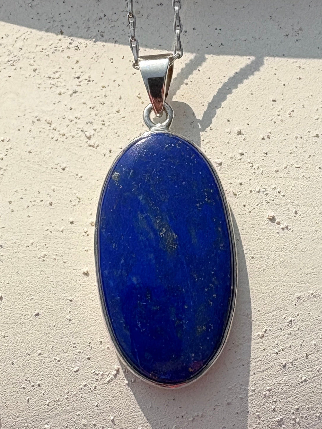 A silver chain necklace with an oval blue lapis lazuli pendant, displayed on a white speckled ceramic pitcher outdoors.