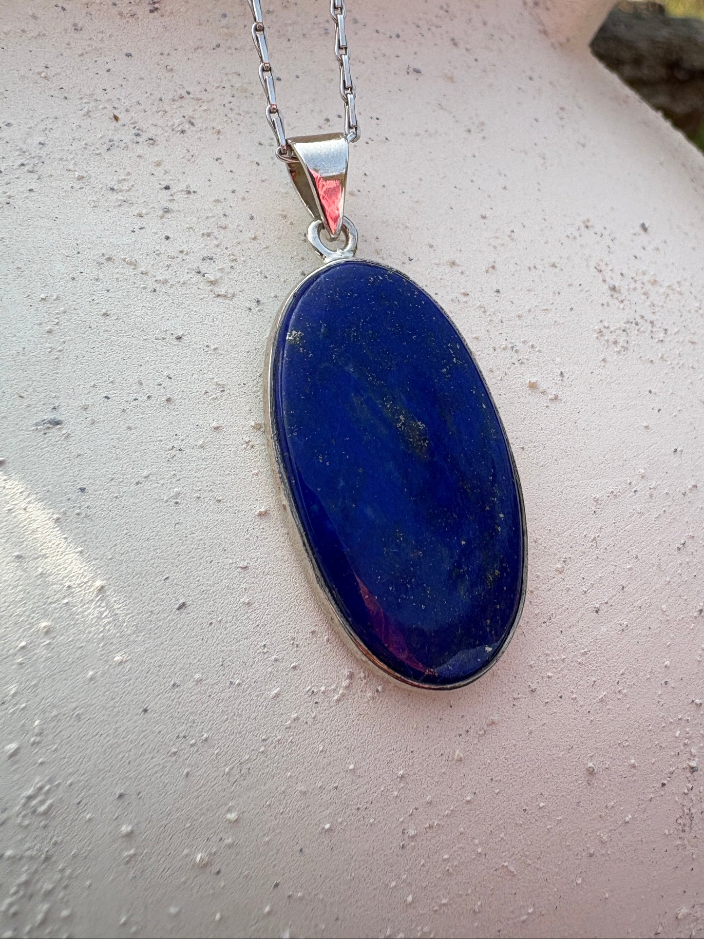 A silver chain necklace with a polished oval dark blue pendant, displayed on a textured white ceramic pitcher outdoors.