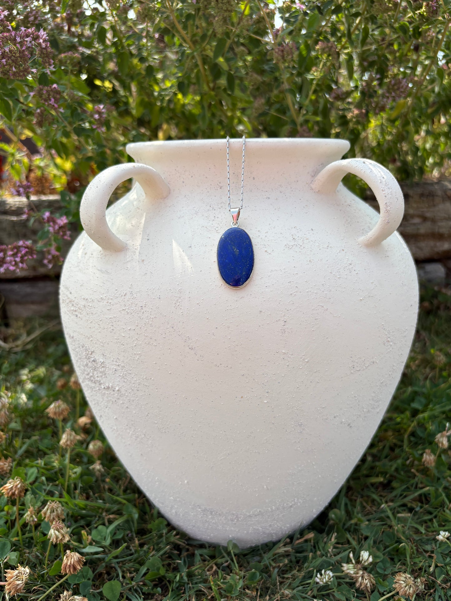 A white ceramic vase with two loop handles and a small oval blue pendant on a silver chain, photographed outdoors on grass with flowers.