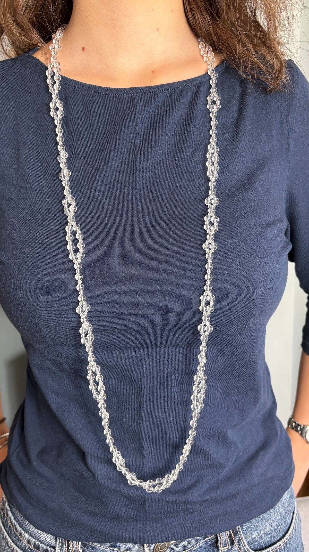 A woman wearing a navy blue three-quarter sleeve top and blue jeans, wearing a long tantric necklace. 