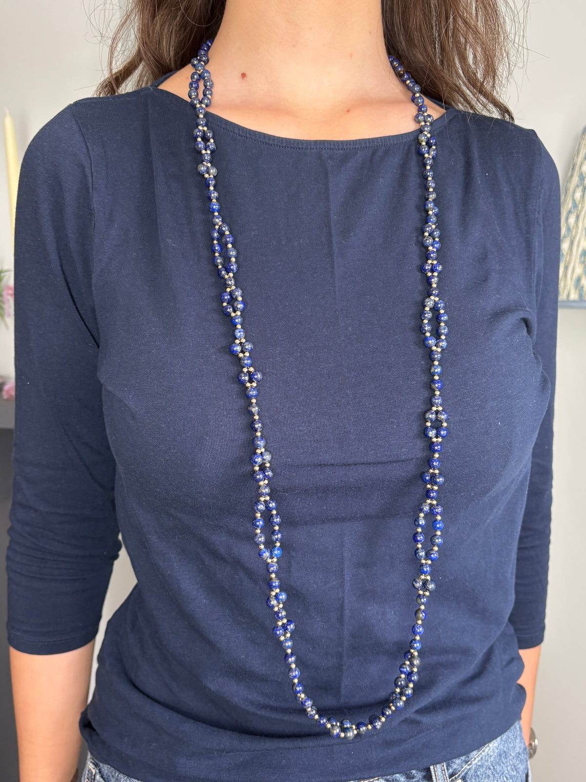 A woman wearing a navy blue long-sleeve top and blue jeans, styled with a long tantric beaded necklace featuring lapis lazuli beads.