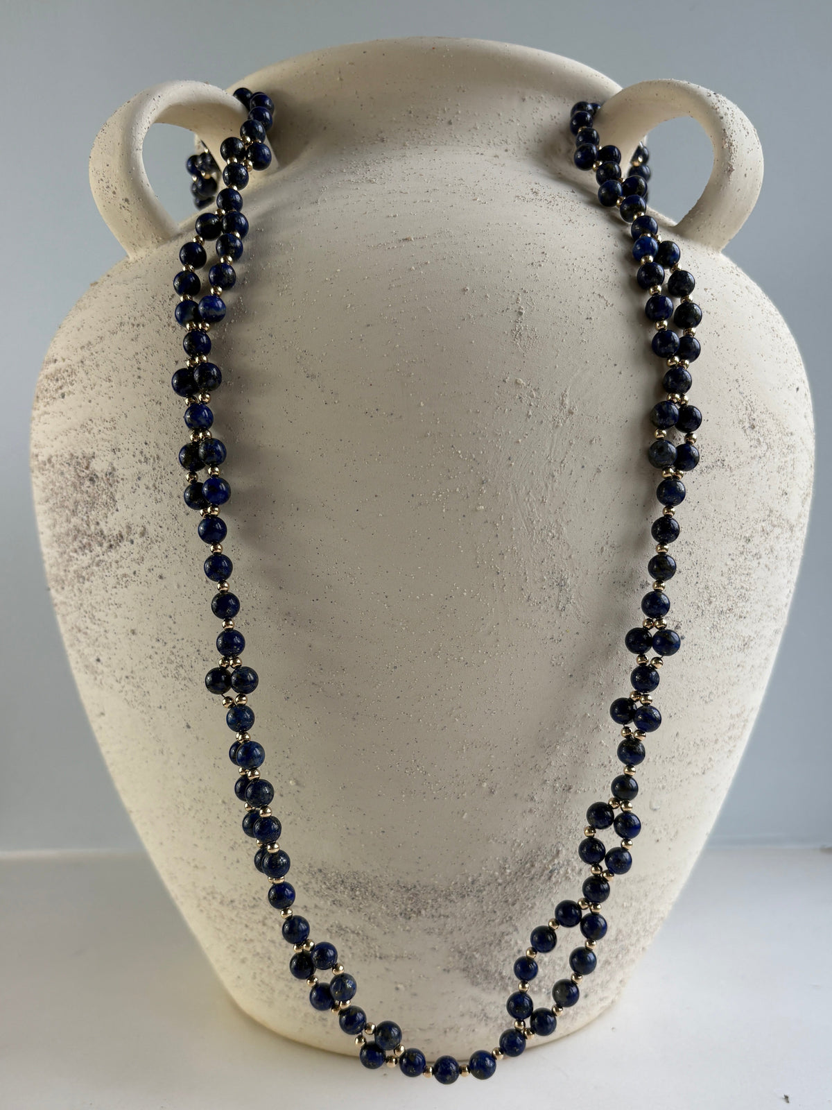 A long tantric beaded necklace made of dark lapis lazuli beads draped over a textured off-white ceramic vase display.