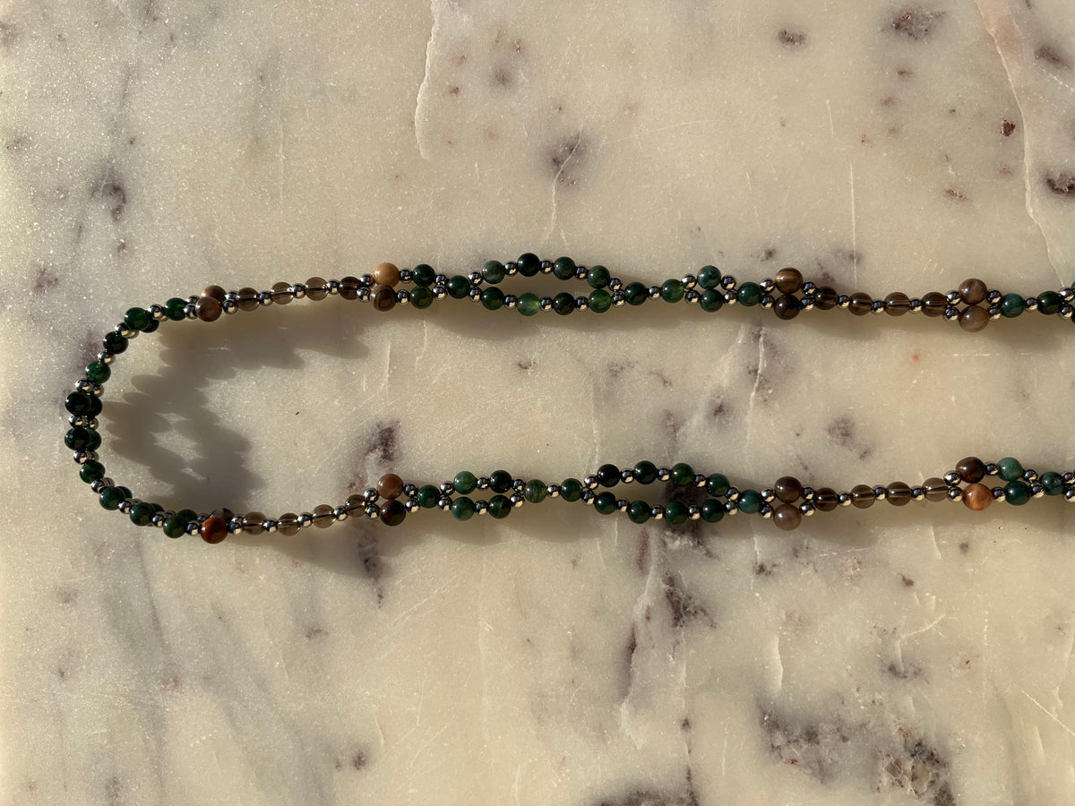 A beaded necklace made of dark green, brown, and metallic silver beads, laid out in a U-shape on a light beige marble surface.