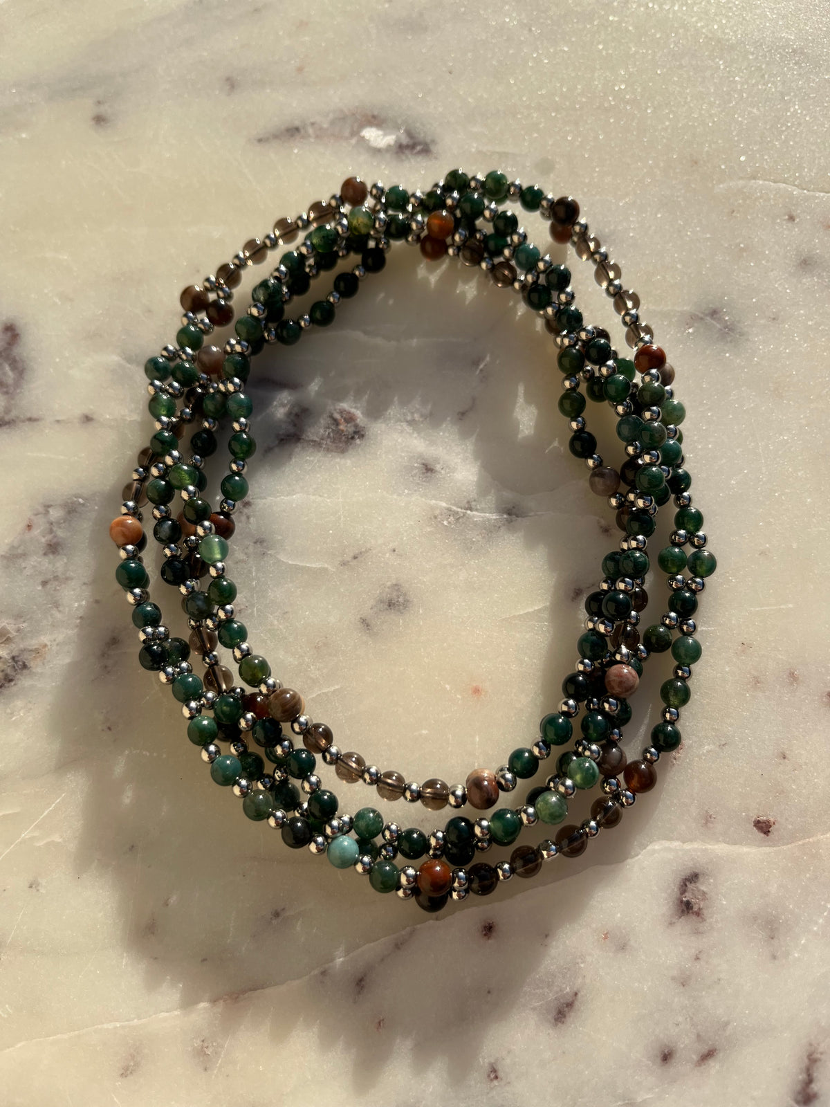 A multi-strand beaded wrap bracelet featuring green, brown, and gray gemstone beads, displayed on a light marble surface.
