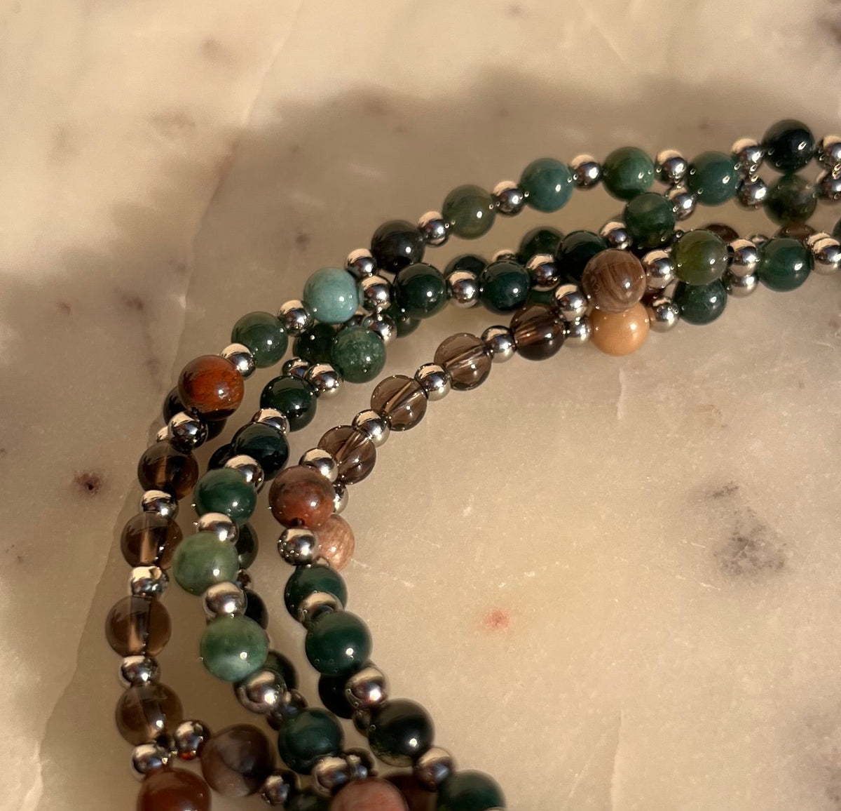 A close-up of a multi-strand beaded necklace with green, brown, and reddish stones separated by small silver beads, resting on a white marble surface.