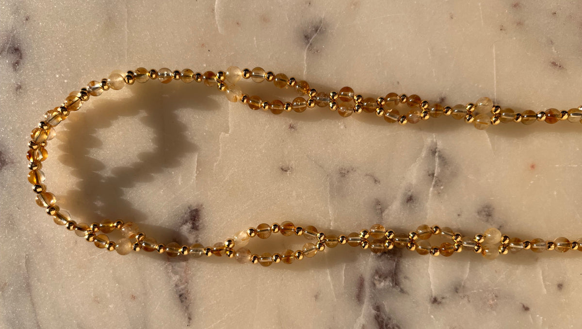 A delicate gold chain necklace with small faceted beads, laid out on a light beige marble surface.