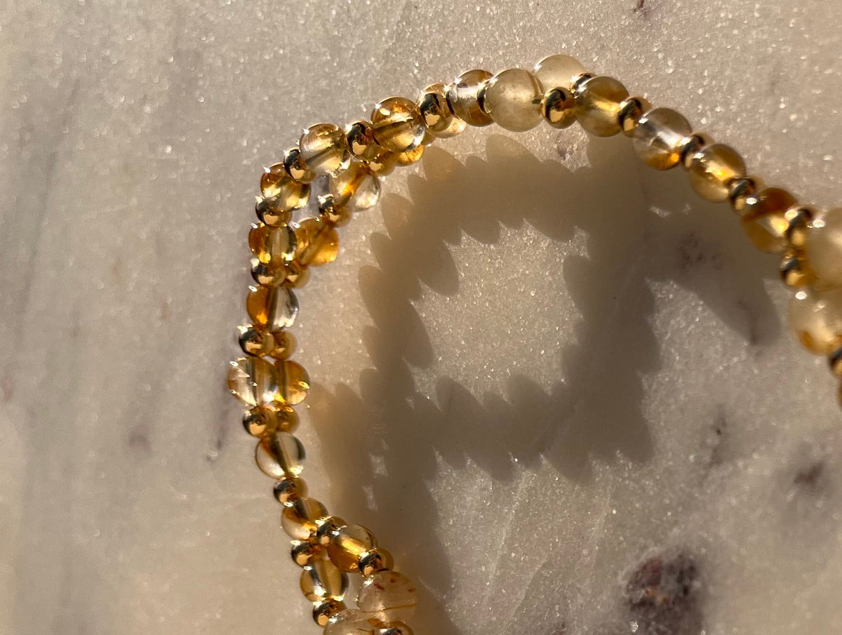 A close-up of a delicate beaded chain necklace with small amber-brown beads and translucent white beads, arranged on a white marble surface.
