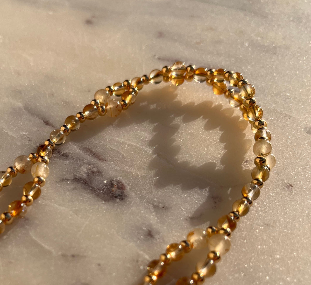 A close-up of a gold-toned beaded chain necklace draped on a white marble surface with gray veining.