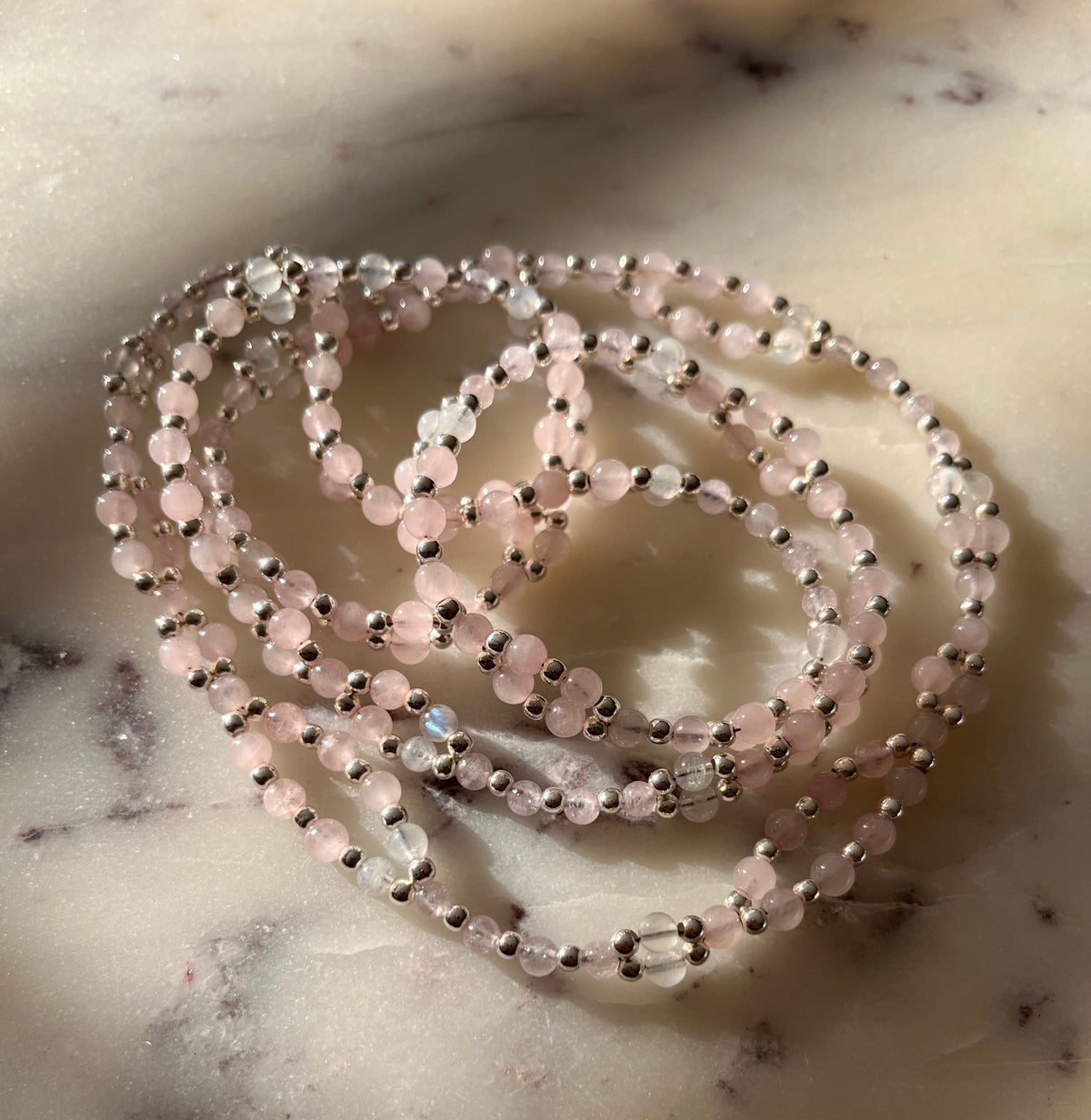 A coiled strand of beaded jewelry made of small pale pink beads separated by metallic silver beads, resting on a light marble surface.