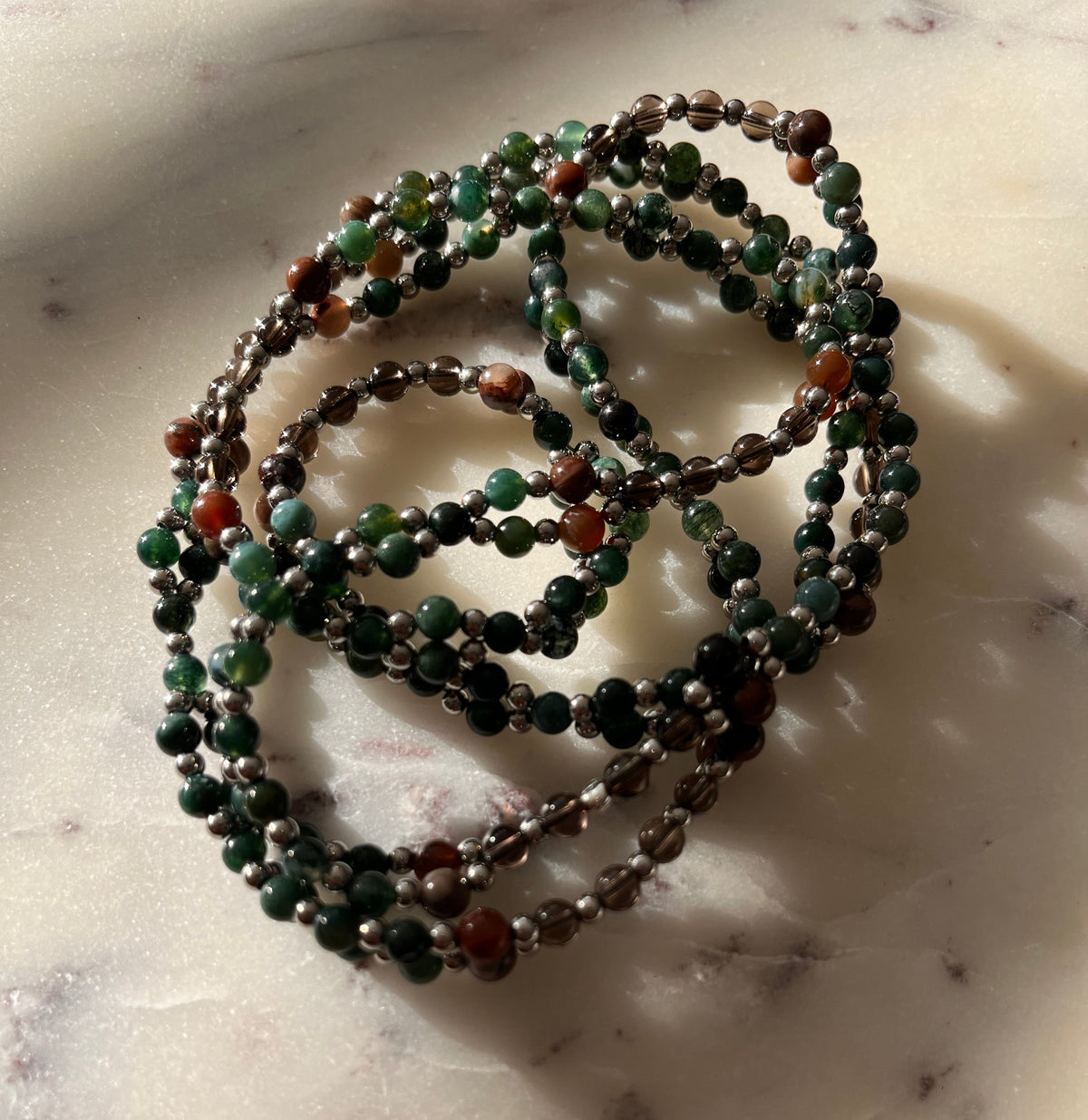 A coiled beaded necklace or bracelet made of small round beads in dark green, black, brown, and reddish-orange, resting on a light marble-like surface with soft shadows.