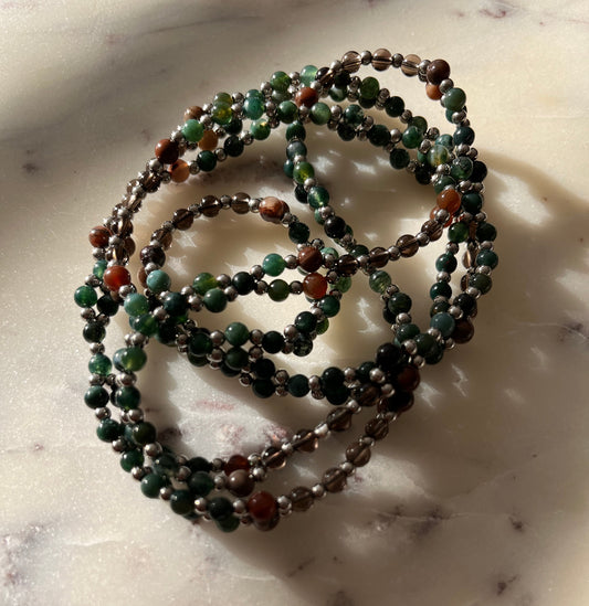 A coiled beaded necklace or bracelet made of small round beads in dark green, black, brown, and reddish-orange, resting on a light marble-like surface with soft shadows.