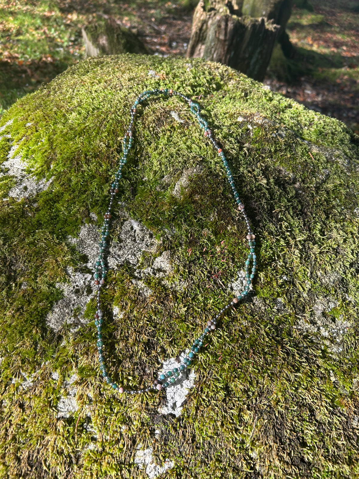 A beaded necklace with dark green and black beads, accented with small red and blue beads, laid out on a moss-covered rock in an outdoor forest setting.