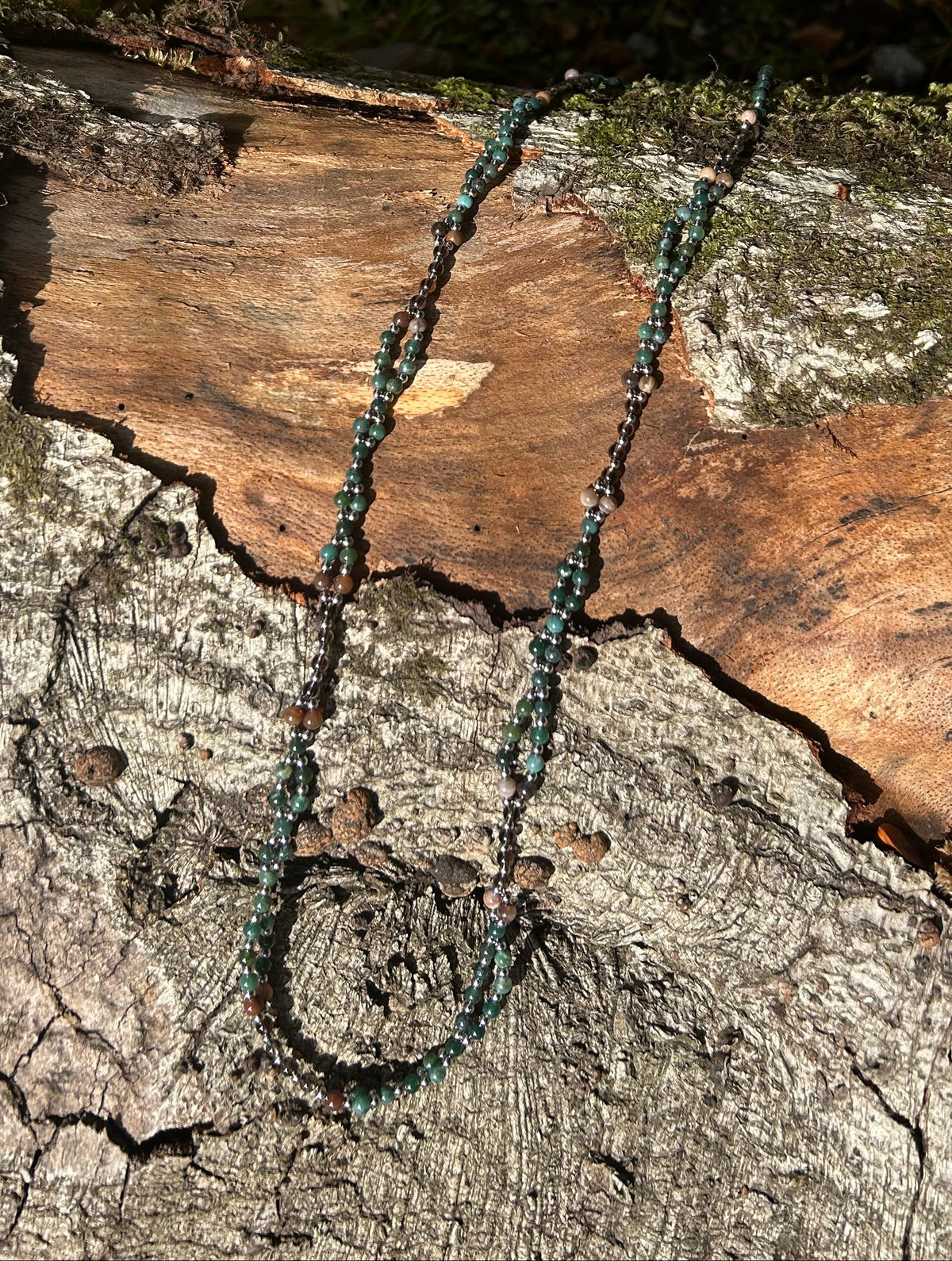 A beaded necklace with dark green and black beads draped over a textured tree log outdoors, with grass and fallen leaves in the background.