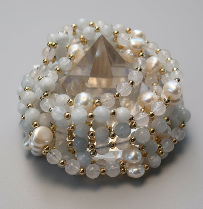 A beaded crystal pyramid display stand featuring celestite and white pearls, moonstone beads, and small gold spacer beads, set against a neutral beige textured fabric background.