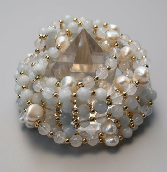 A beaded crystal pyramid display stand featuring celestite and white pearls, moonstone beads, and small gold spacer beads, set against a neutral beige textured fabric background.