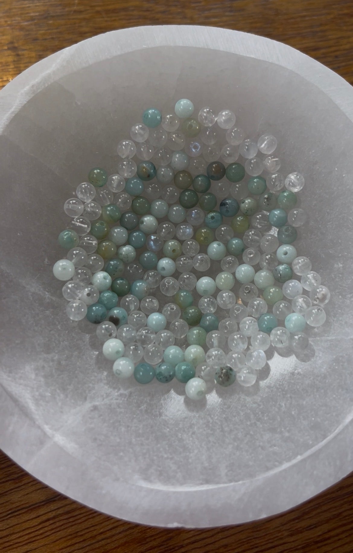 Amazonite and Rainbow Moonstone beads resting in a carved selenite bowl before being handcrafted into a sacred geometric tantric necklace, photographed in natural light.