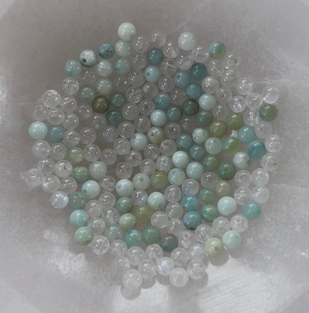 Amazonite and Rainbow Moonstone beads resting in a carved selenite bowl before being handcrafted into a sacred geometric tantric necklace, photographed in natural light.